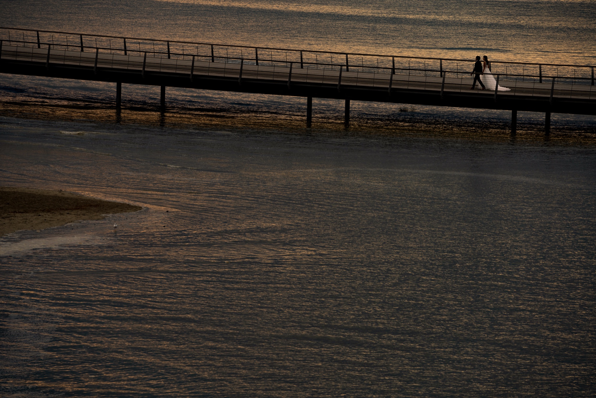 Bride and groom walk hand in hand on the Mont-Saint-Michel footbridge above the water, silhouettes on the sunset reflections — after-wedding session in Normandy — Arnaud Chapelle