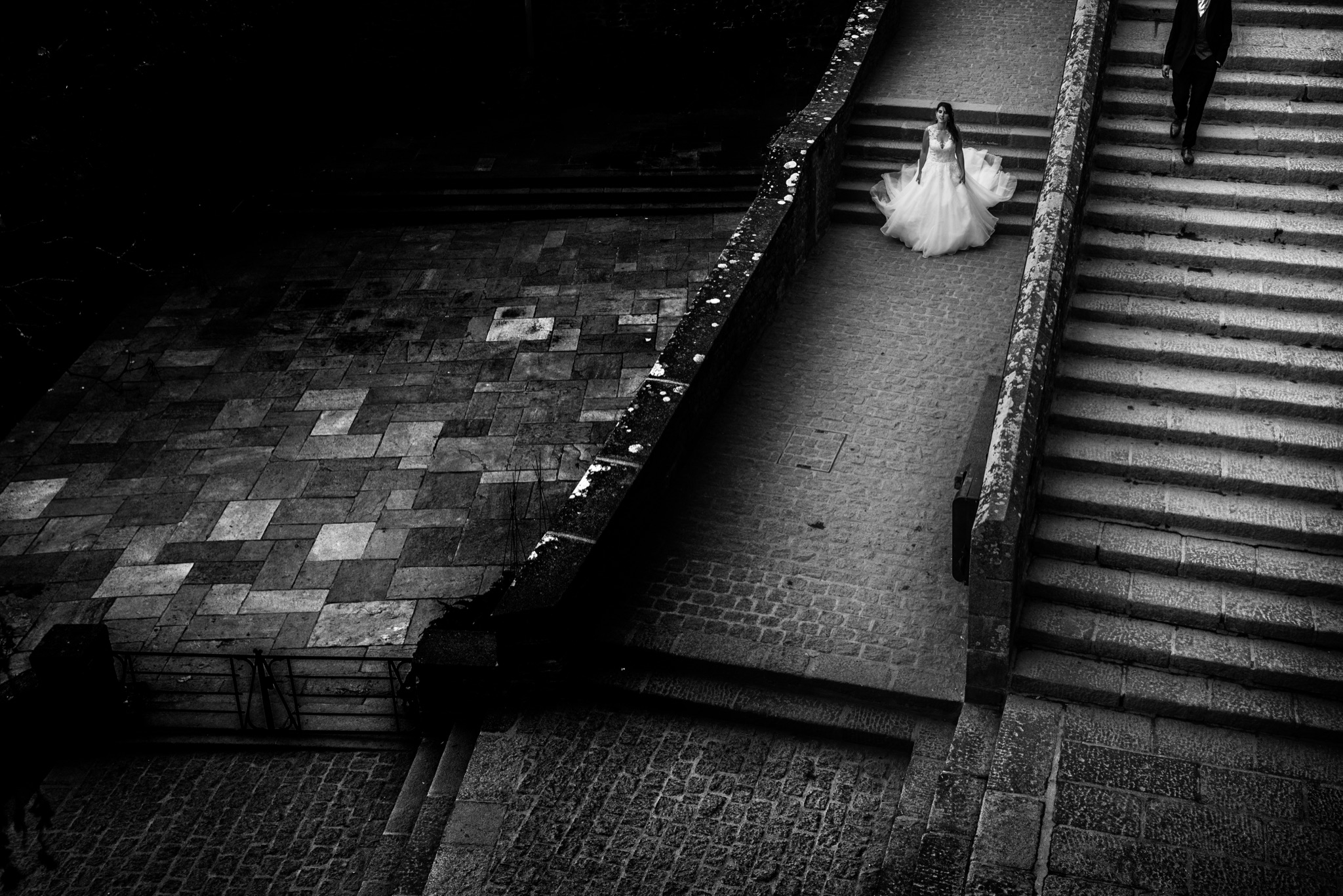 The bride spreads her gown at the foot of the grand staircase of Mont-Saint-Michel while the groom walks down the steps above, in black and white — Arnaud Chapelle