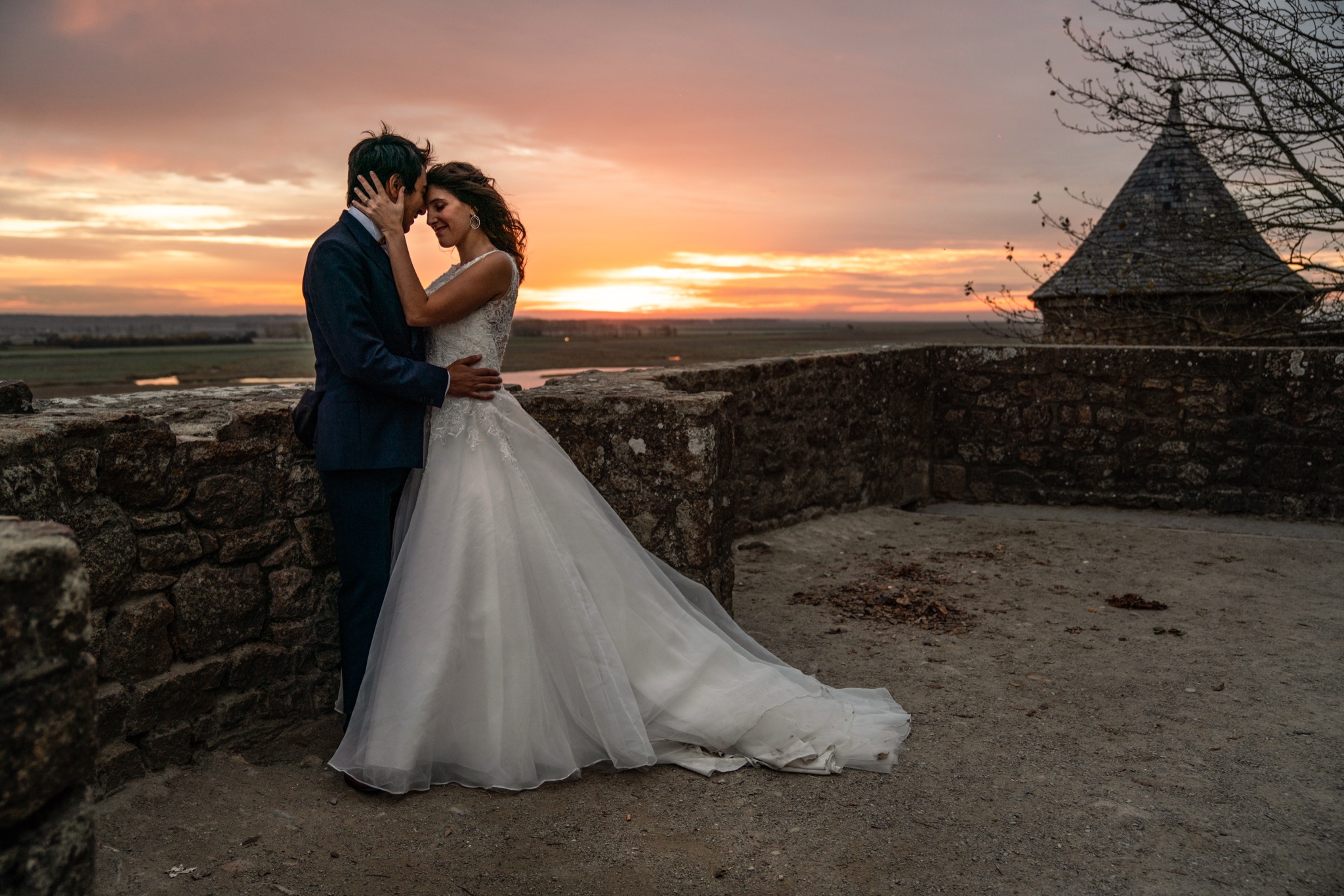 Bride and groom forehead to forehead on a rampart terrace of Mont-Saint-Michel, the turret and the bay behind them at sunset — Arnaud Chapelle