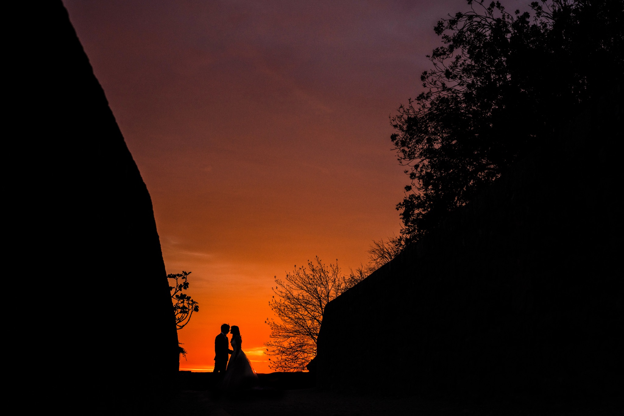 Silhouettes of the couple kissing between the dark masses of the Mont-Saint-Michel ramparts, against a blazing orange and purple sky — Arnaud Chapelle