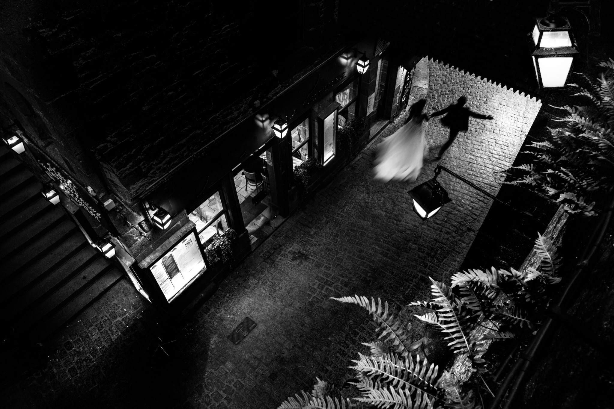 The couple runs through a cobbled lane of Mont-Saint-Michel at night, between lanterns and the lit front of a restaurant, in black and white — Arnaud Chapelle