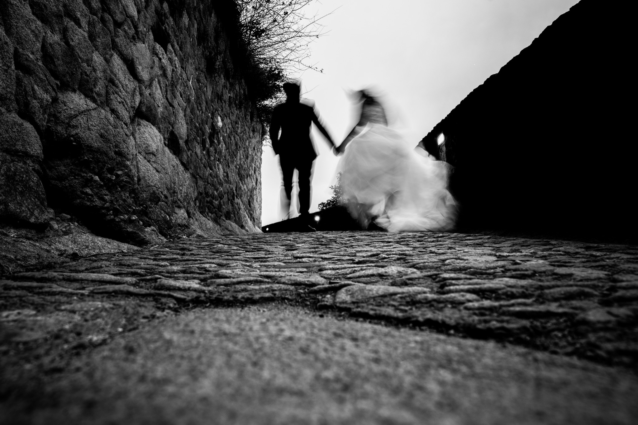 At cobblestone level, the couple walks away hand in hand through a lane of Mont-Saint-Michel, the dress in motion, in black and white — Arnaud Chapelle
