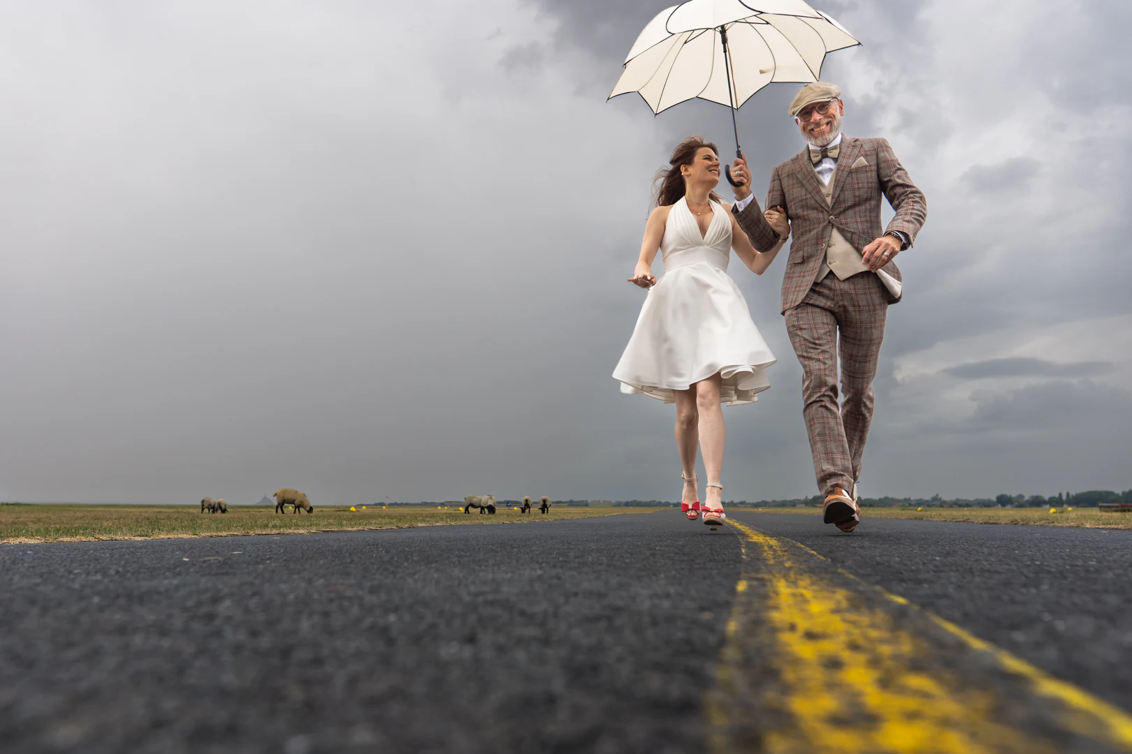Les mariés remontent l'axe de la piste sous un parapluie blanc, ciel d'orage, moutons en pâture sur les herbus de part et d'autre — after-wedding à l'aérodrome d'Avranches — Arnaud Chapelle