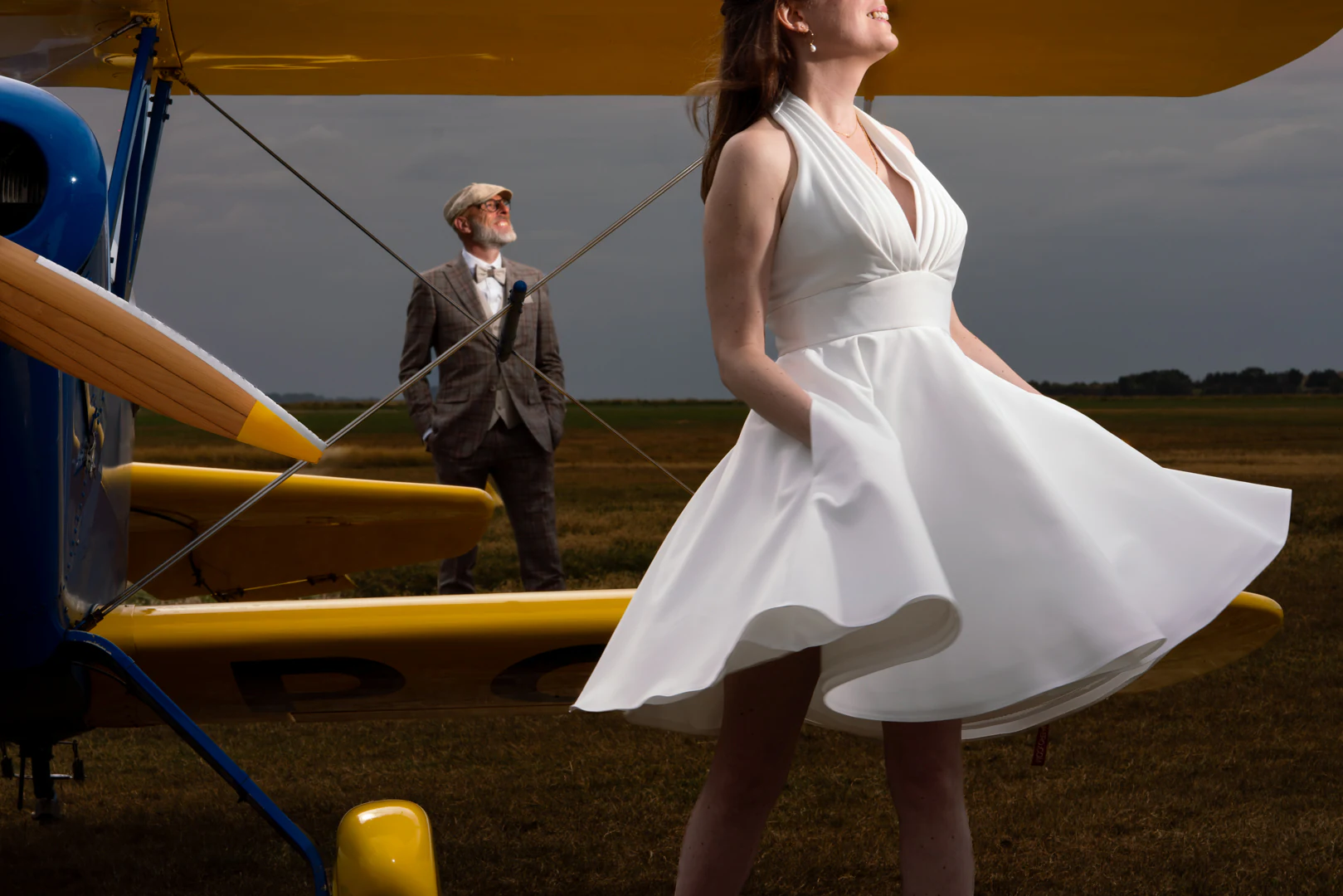 La mariée fait tourner sa robe courte sous l'aile jaune d'un biplan, le marié en costume à carreaux et casquette plate la regarde en arrière-plan — after-wedding à l'aérodrome d'Avranches — Arnaud Chapelle