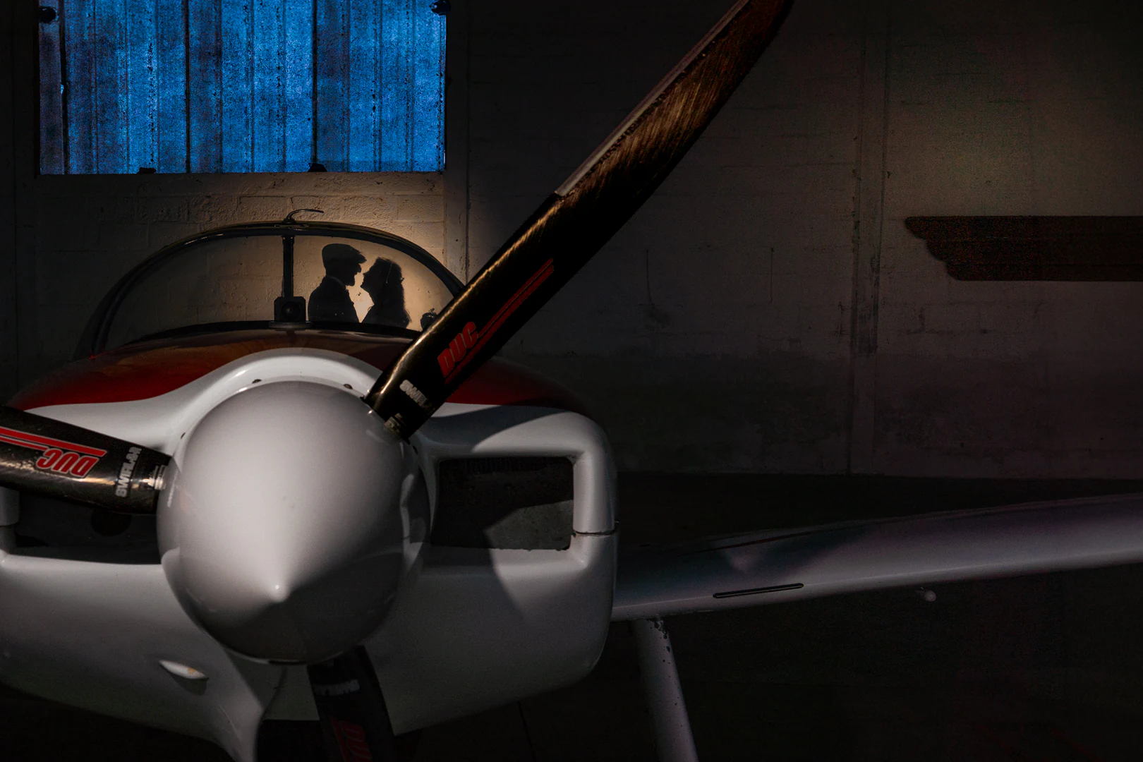 Dans un hangar, la silhouette des mariés front contre front à travers la verrière d'un avion, une lucarne bleue au-dessus, l'hélice au premier plan — after-wedding à l'aérodrome d'Avranches — Arnaud Chapelle