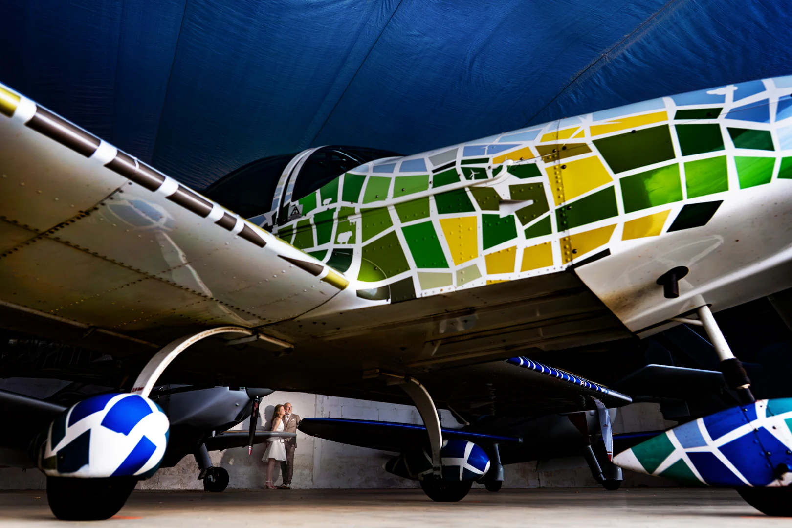 Sous les ailes d'avions aux livrées colorées alignés dans un hangar, les mariés minuscules au fond contre le mur — after-wedding à l'aérodrome d'Avranches — Arnaud Chapelle