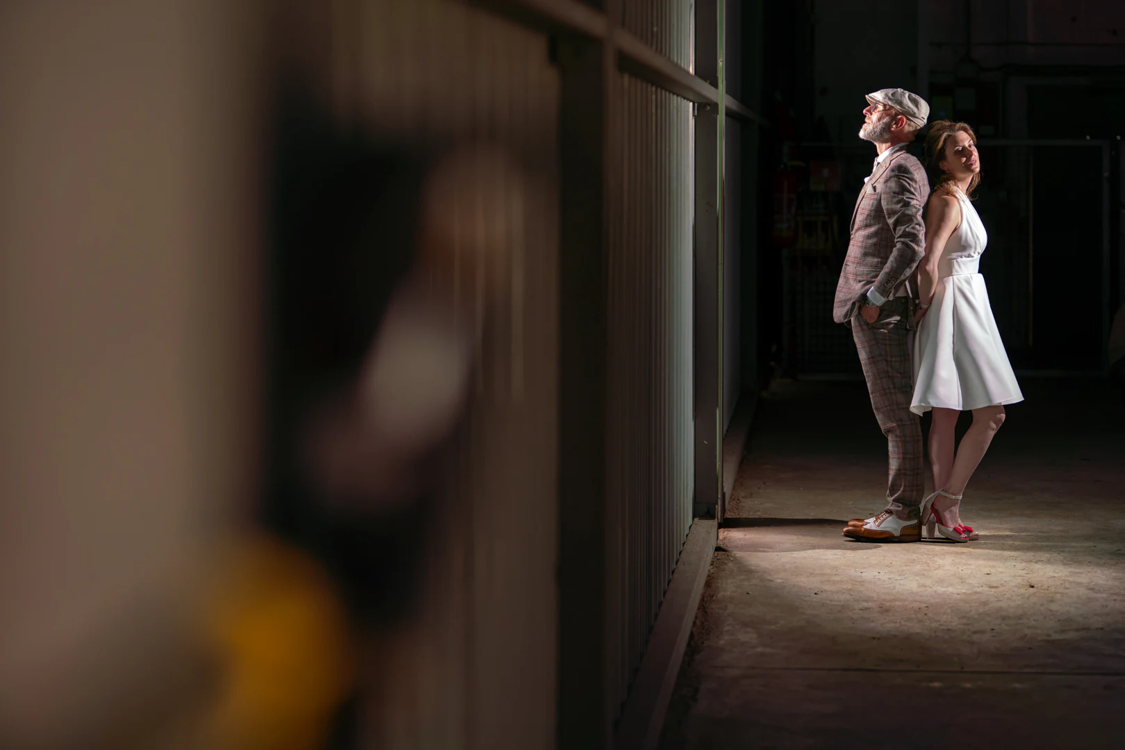 Les mariés adossés dos à dos contre la paroi d'un hangar, éclairés par la lumière qui entre par la porte entrouverte — after-wedding à l'aérodrome d'Avranches — Arnaud Chapelle