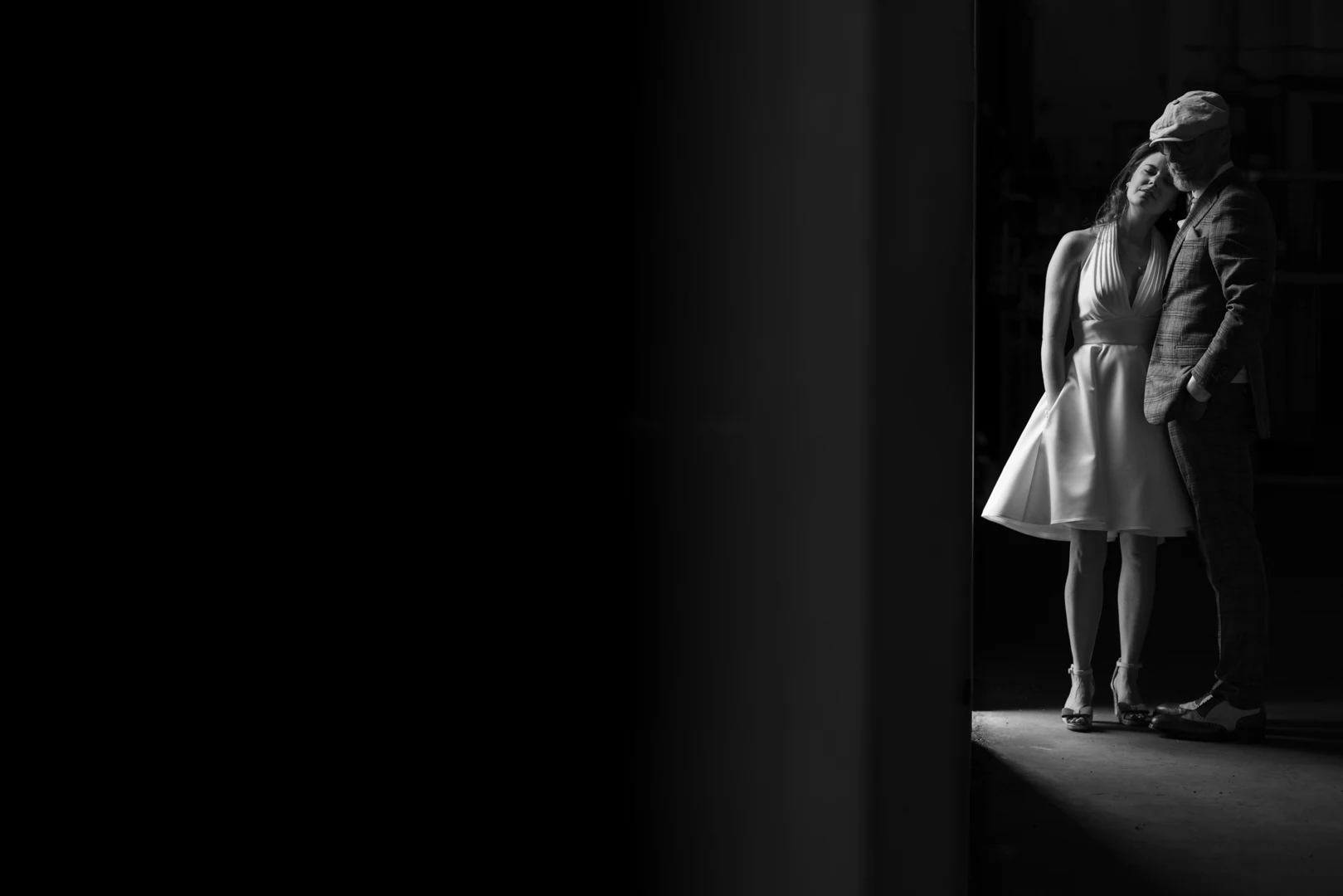En noir et blanc, les mariés serrés l'un contre l'autre dans un rai de lumière au bord d'un hangar, le reste du cadre dans le noir — after-wedding à l'aérodrome d'Avranches — Arnaud Chapelle
