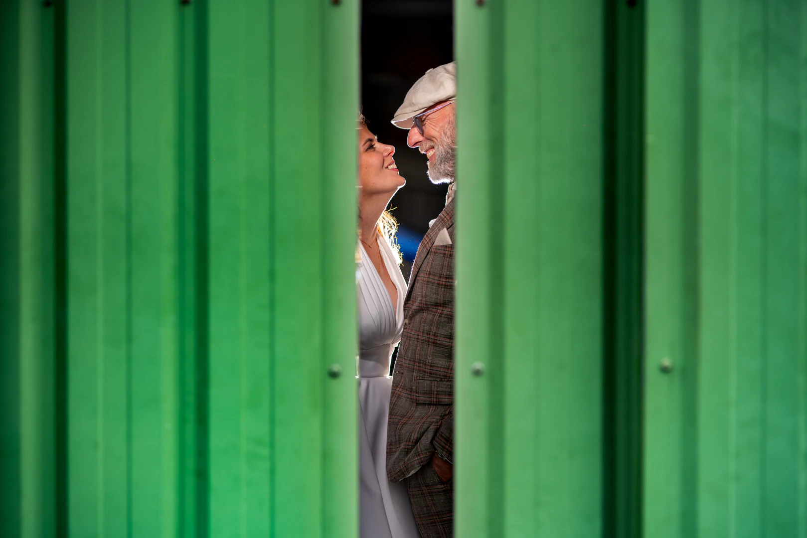 Les mariés se regardent, aperçus par l'entrebâillement de deux portes de hangar vertes — after-wedding à l'aérodrome d'Avranches — Arnaud Chapelle