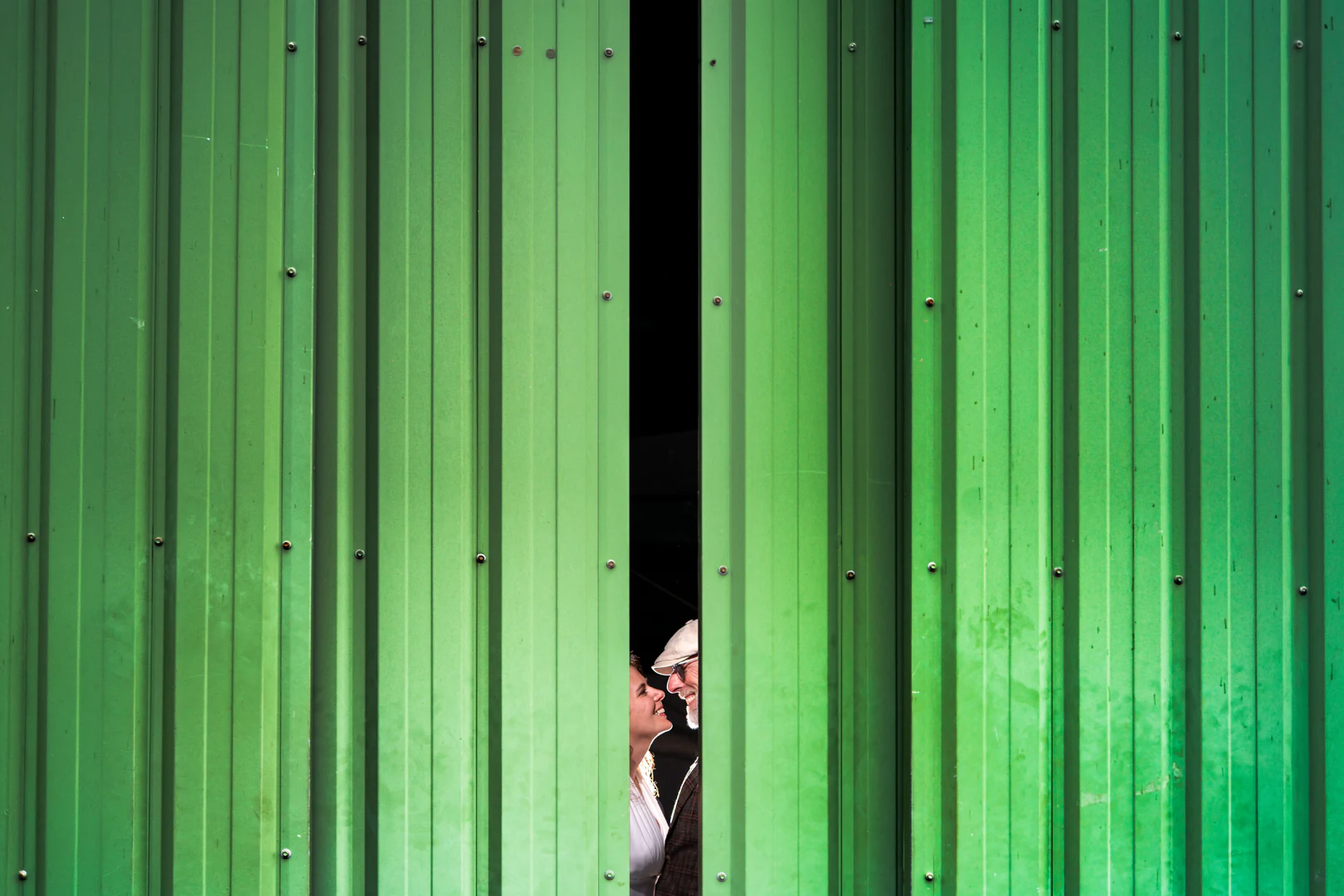 Large mur de tôle verte d'un hangar d'aérodrome, les mariés visibles dans la fente étroite entre les deux portes — after-wedding à l'aérodrome d'Avranches — Arnaud Chapelle