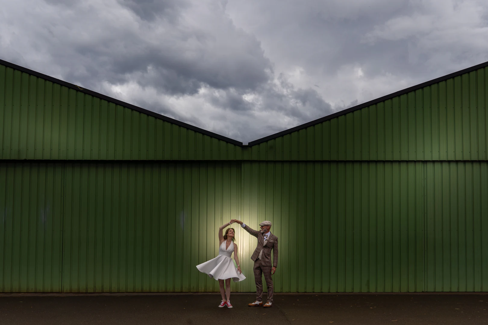 Le marié fait tourner la mariée devant les portes vertes d'un hangar sous un ciel d'orage, elle porte des baskets rouges — after-wedding à l'aérodrome d'Avranches — Arnaud Chapelle