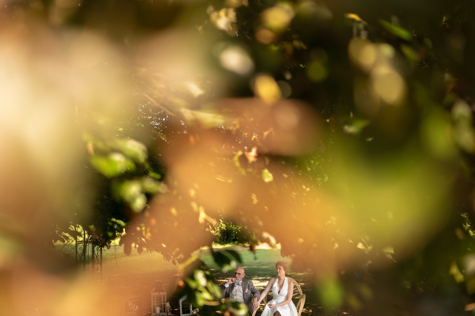 Charlotte et Laurent assis dans le parc, aperçus à travers le feuillage — mariage aux Châteaux de La Lucerne d'Outremer — Arnaud Chapelle
