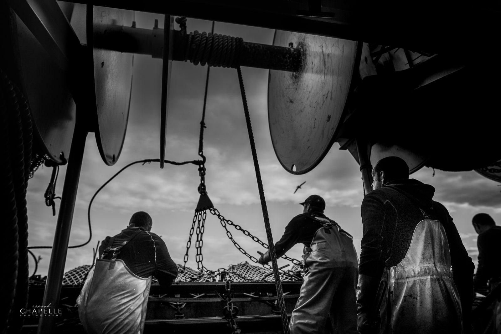 Les marins sous les dragues, silhouettes à contre-jour sur le pont, en noir et blanc — Télémaque, Grandcamp-Maisy — Arnaud Chapelle