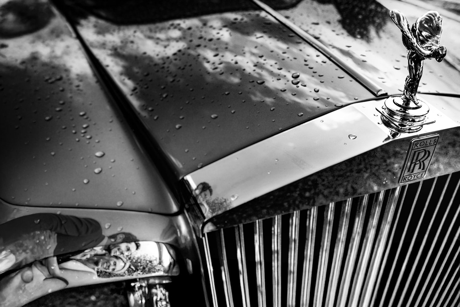 Raindrops on the bonnet of a Rolls-Royce, the couple reflected in the chrome — wedding photograph by Arnaud Chapelle