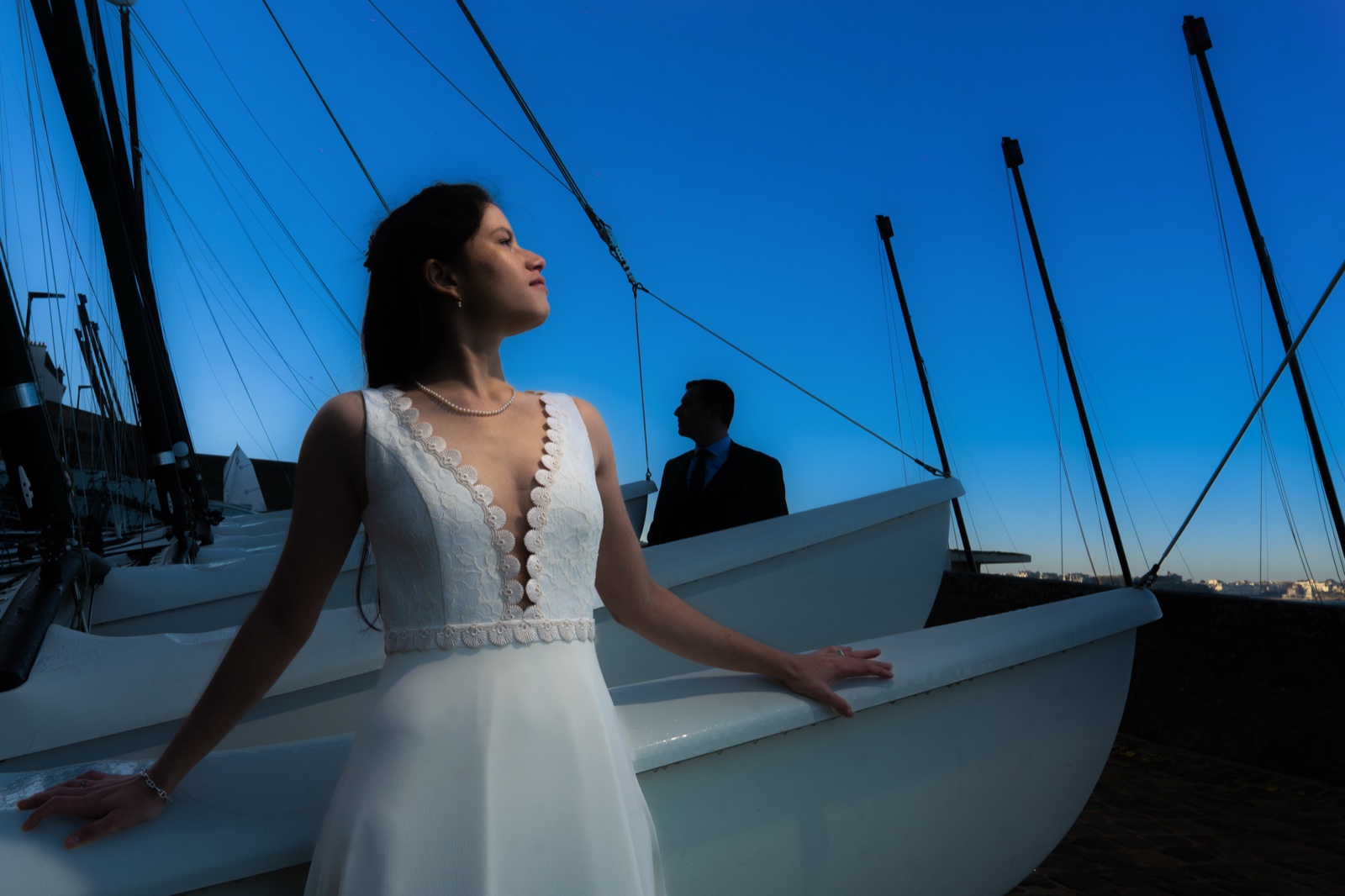 Marine en robe de mariée appuyée à la coque d'un doris à l'heure bleue, Alexandre en arrière-plan — Saint-Malo — Arnaud Chapelle