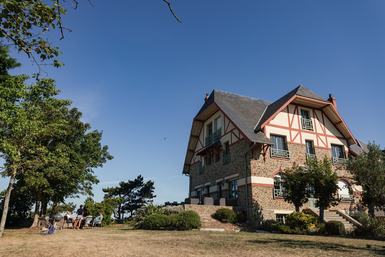 La maison de pierre et ses colombages, les premiers invités installés à l'ombre dans le jardin — mariage à Granville — Arnaud Chapelle
