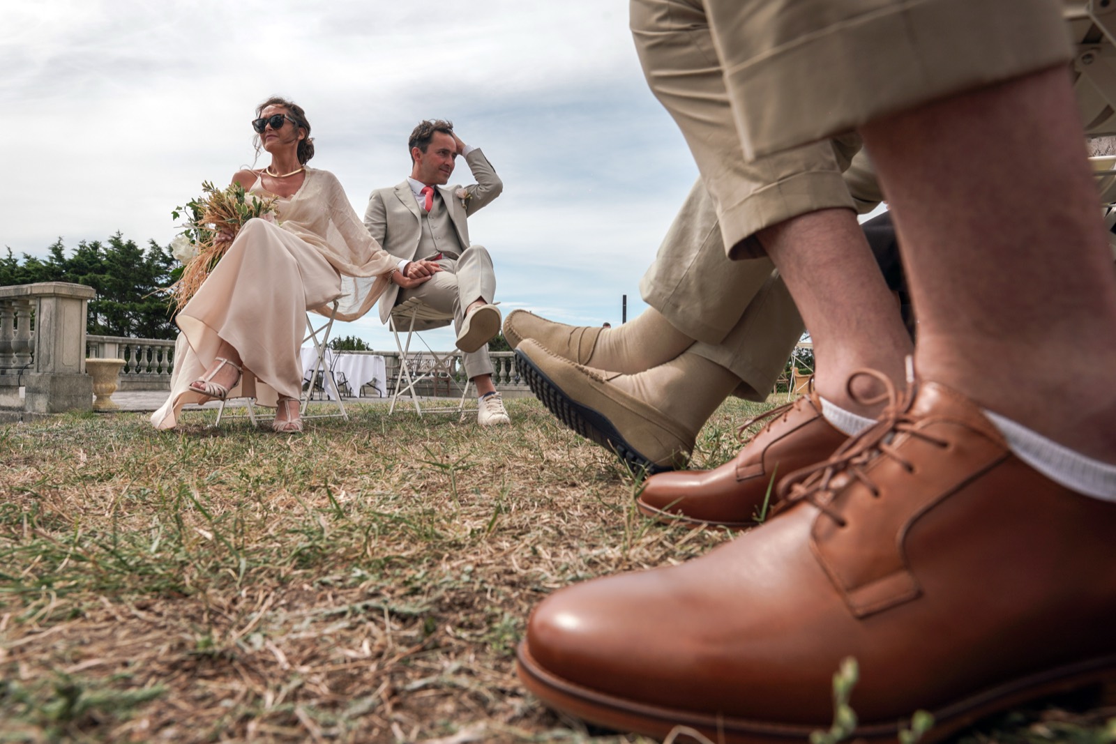 Les mariés assis main dans la main pendant la cérémonie, vus derrière les chaussures d'un invité — mariage au Château de la Crête, Granville — Arnaud Chapelle