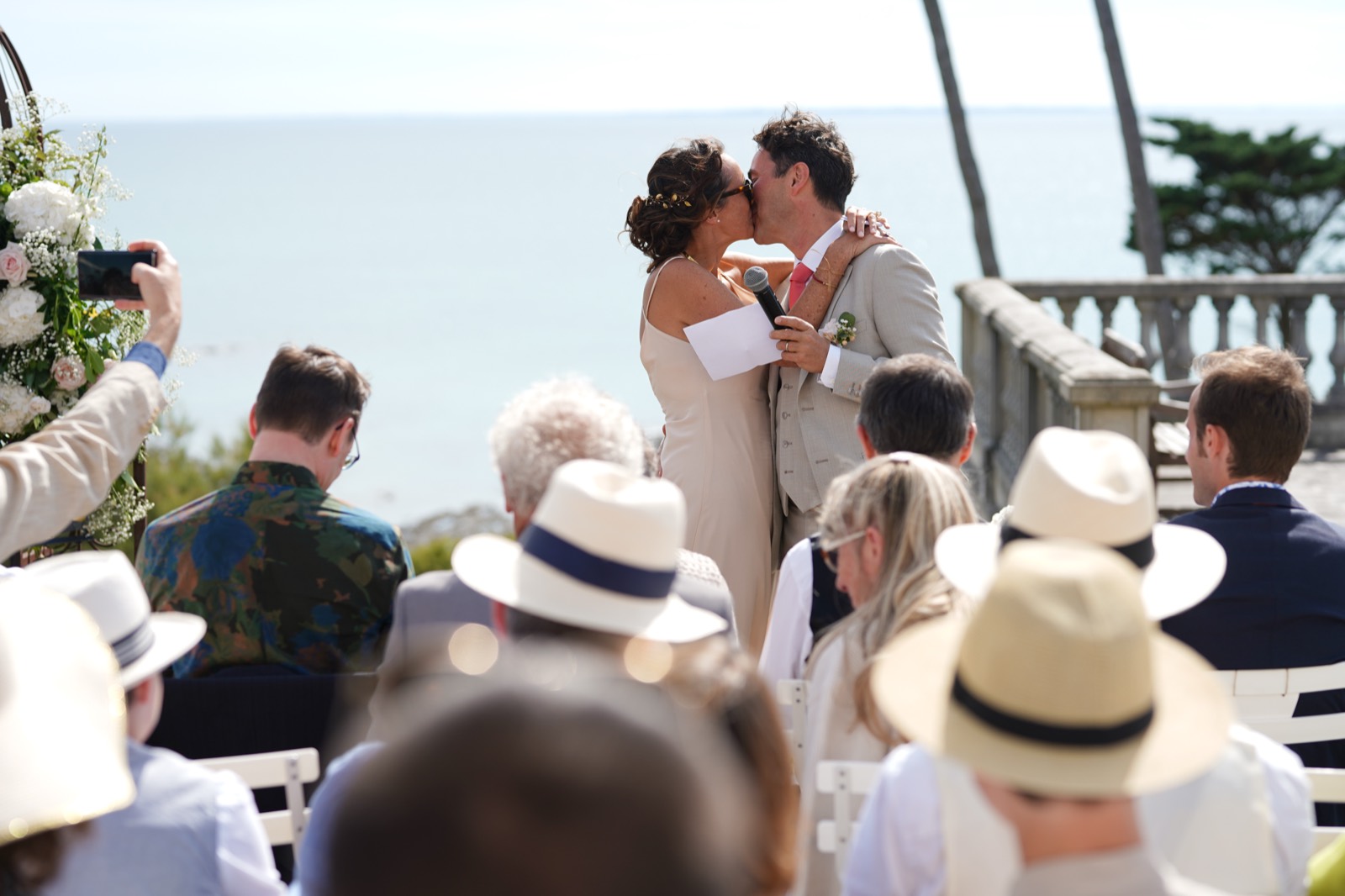 Le baiser des mariés face à la mer, au milieu des chapeaux panama des invités — mariage au Château de la Crête à Granville (Manche) — Arnaud Chapelle
