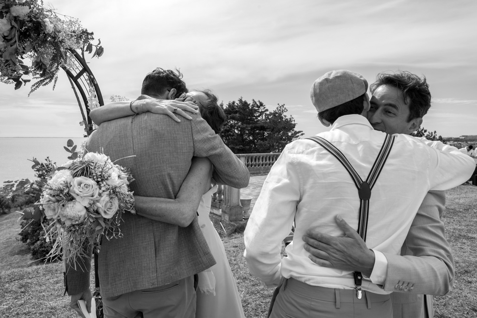 Les embrassades juste après la cérémonie, en noir et blanc, devant l'arche fleurie et la mer — mariage à Granville — Arnaud Chapelle