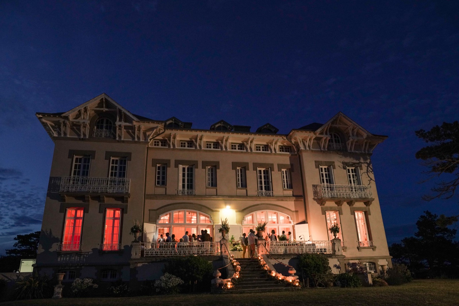 La façade du Château de la Crête éclairée à la nuit tombée, bougies le long de l'escalier et invités sur la terrasse — mariage à Granville — Arnaud Chapelle