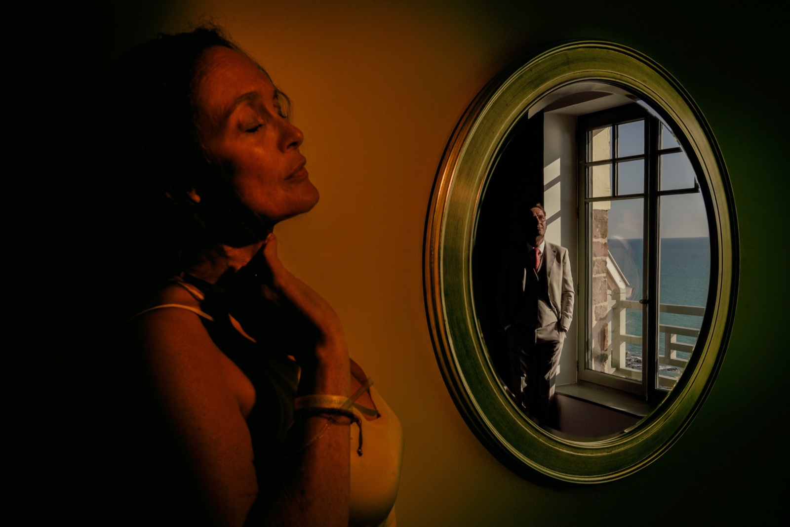 La mariée de profil et le marié reflété dans un miroir ovale devant la fenêtre ouverte sur la mer — after-wedding au Château de la Crête, Granville — Arnaud Chapelle