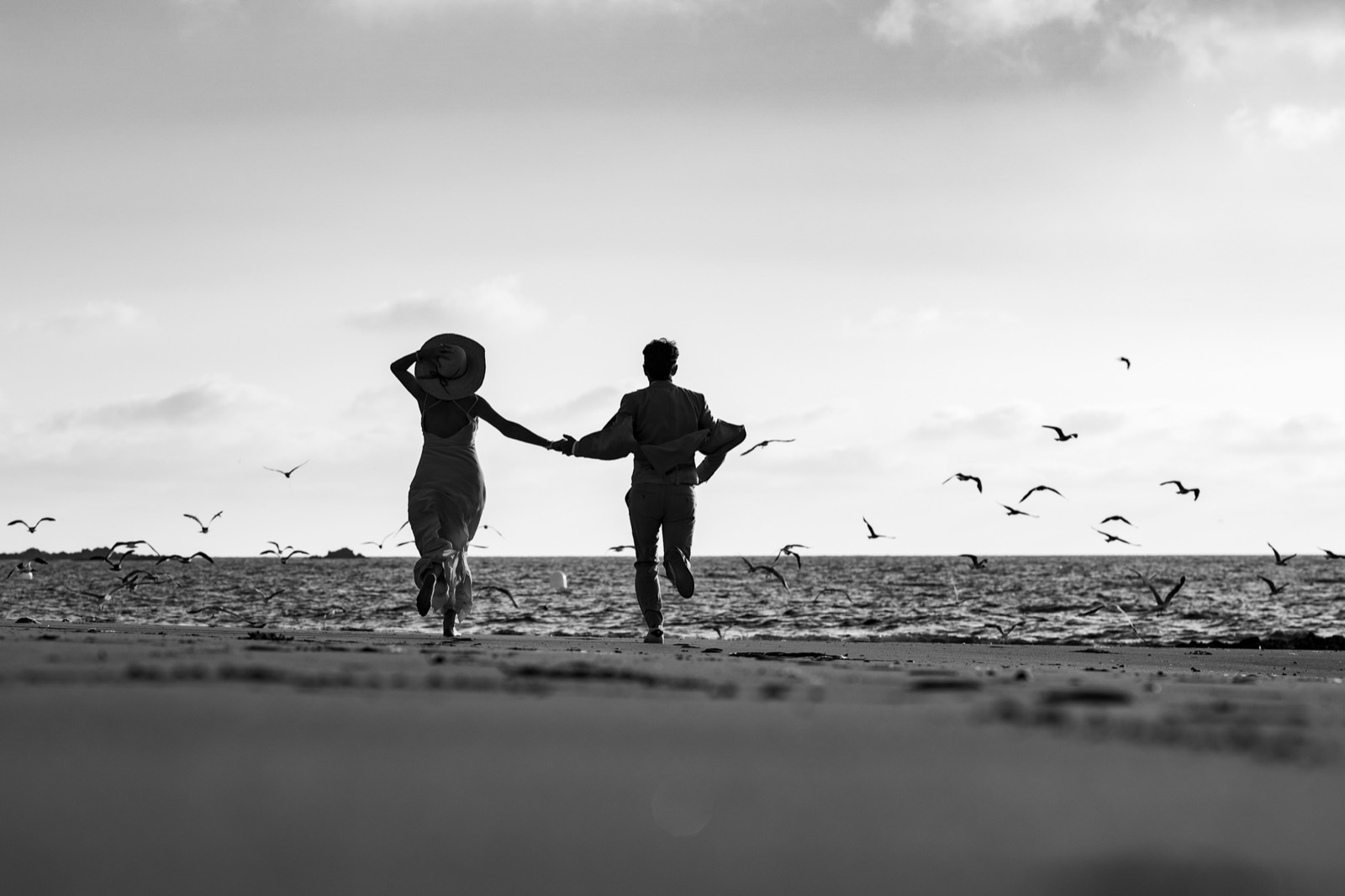 Les mariés courent sur la plage main dans la main et font s'envoler les mouettes, en noir et blanc — séance couple after-wedding à Granville — Arnaud Chapelle