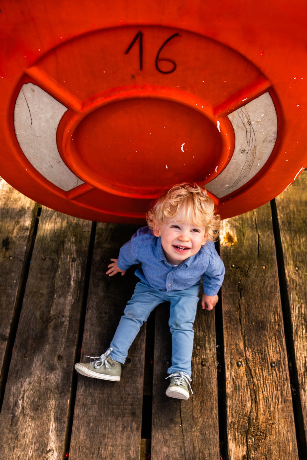 Le petit garçon assis sous une grande bouée orange numérotée 16, sur le ponton en bois — séance famille à Cancale — Arnaud Chapelle