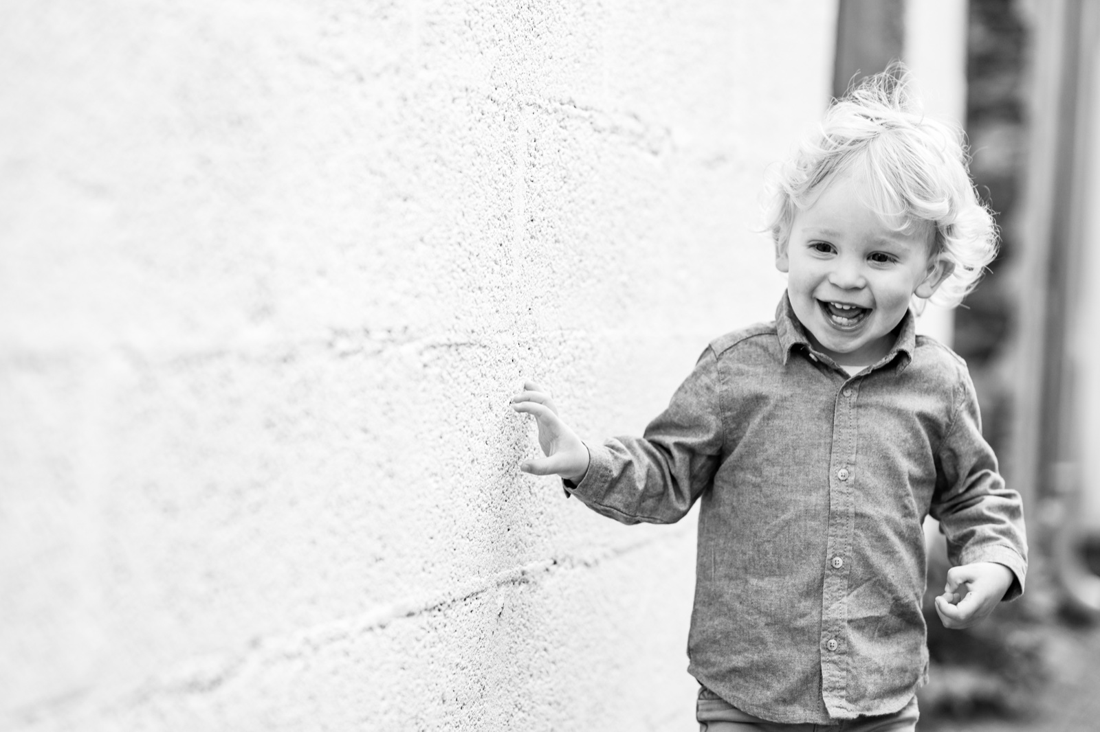 Le petit garçon longe un mur blanc en riant, en noir et blanc — séance famille à Cancale — Arnaud Chapelle