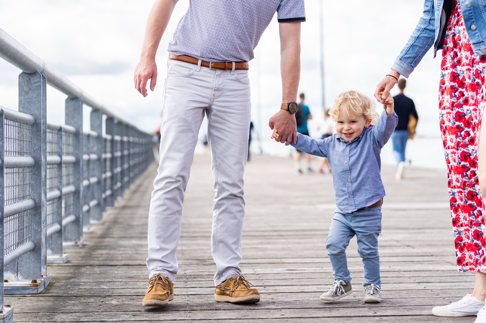 Le petit garçon marche sur le ponton, tenu par les mains de ses parents — séance famille à Cancale — Arnaud Chapelle