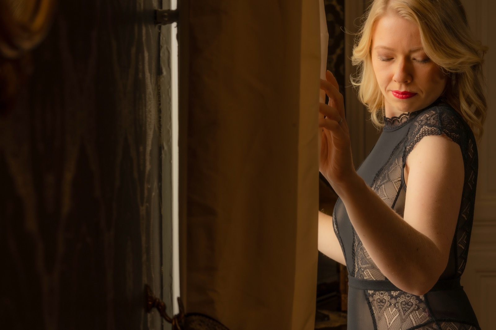 Près de la fenêtre, une main sur le rideau, le regard baissé — séance boudoir près de Bayeux (Calvados) — Arnaud Chapelle