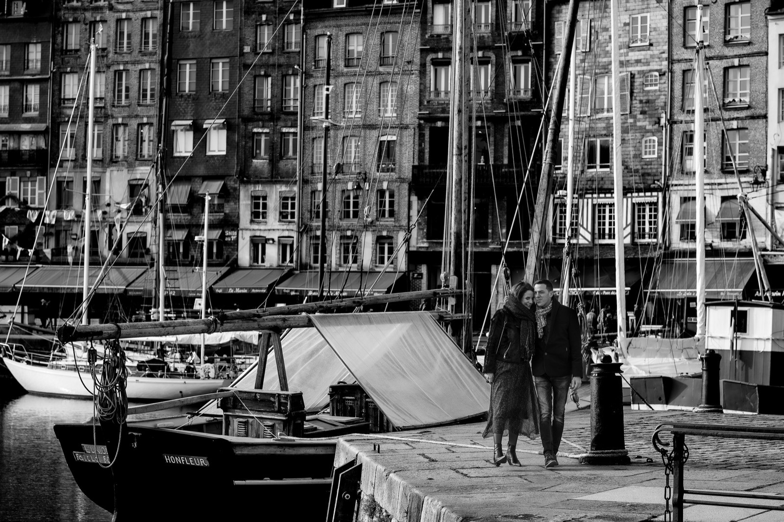 Le couple marche sur le quai du Vieux Bassin, une barque marquée Honfleur au premier plan, en noir et blanc — séance engagement à Honfleur — Arnaud Chapelle
