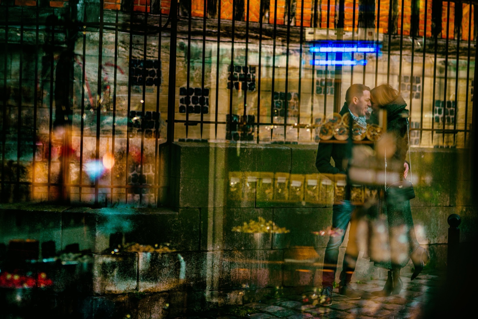 Le couple enlacé derrière les grilles et les reflets d'une vitrine, jeu de superpositions colorées — séance engagement à Honfleur — Arnaud Chapelle