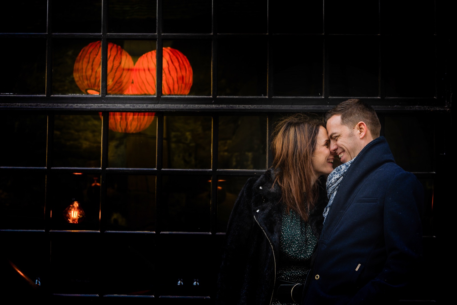Le couple front contre front devant une vitrine noire éclairée de lampions rouges — séance engagement à Honfleur — Arnaud Chapelle