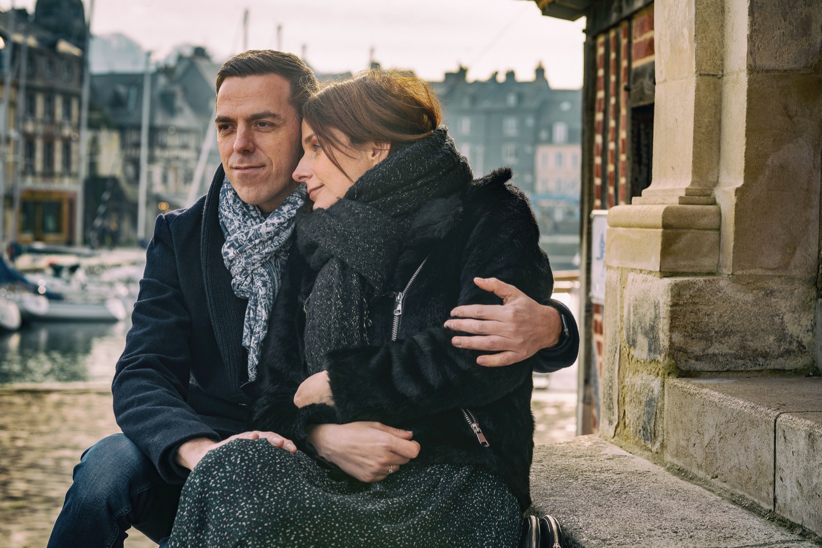 Le couple assis au pied d'un pilier de pierre, elle blottie contre lui, le bassin et les maisons de Honfleur derrière — séance engagement — Arnaud Chapelle