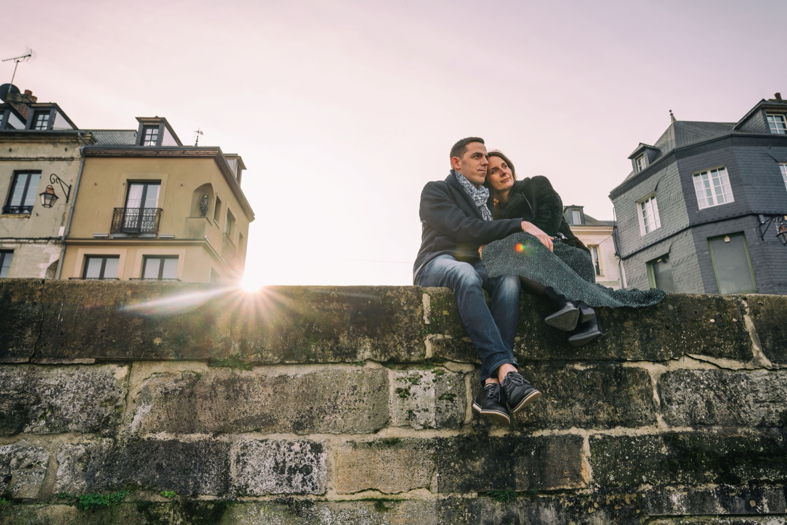 Le couple assis sur un muret de pierre au soleil rasant d'hiver, les maisons d'ardoise de Honfleur en arrière-plan — séance engagement — Arnaud Chapelle