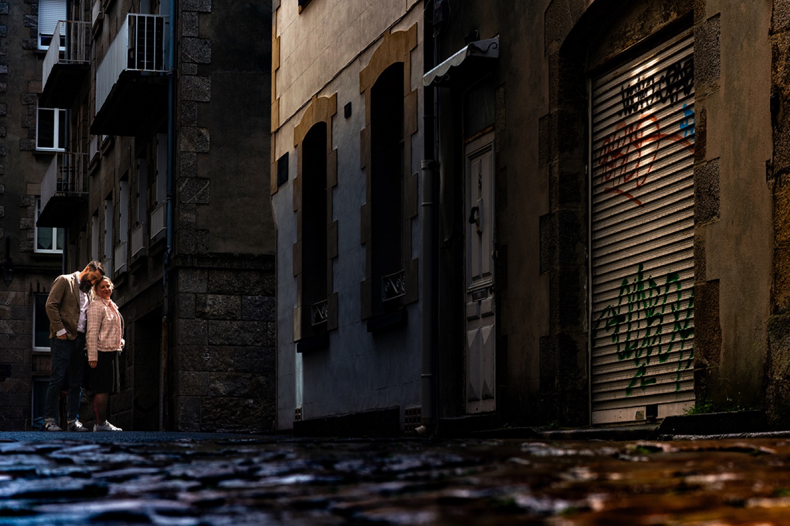 Le couple s'embrasse dans une ruelle pavée de l'intra-muros, façades de granit et rideau tagué — séance engagement à Saint-Malo — Arnaud Chapelle