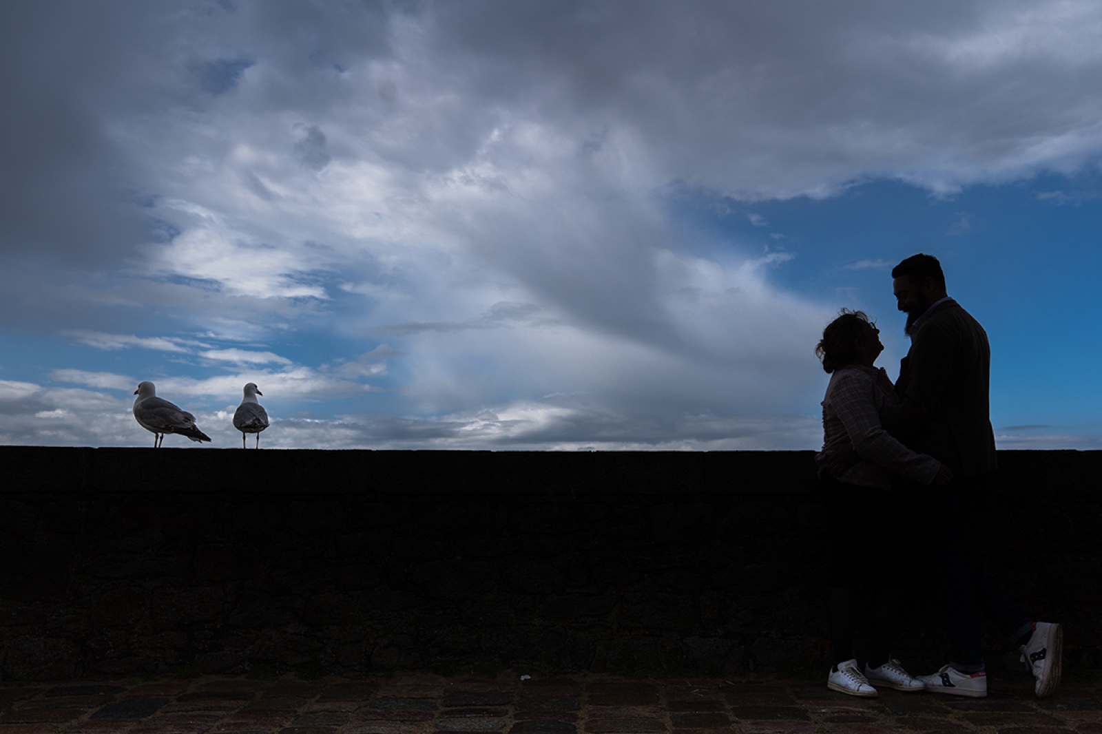 Silhouettes du couple face au ciel, deux goélands posés sur le parapet des remparts — séance engagement à Saint-Malo — Arnaud Chapelle