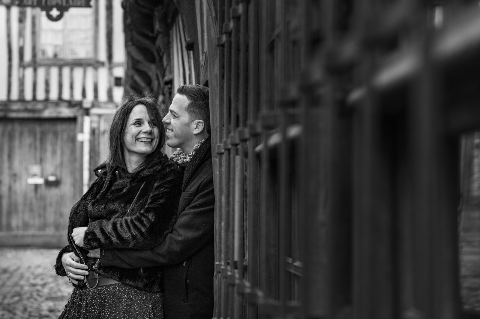 The couple laughs, embracing against a half-timbered facade in an old Honfleur lane, in black and white — engagement session — Arnaud Chapelle