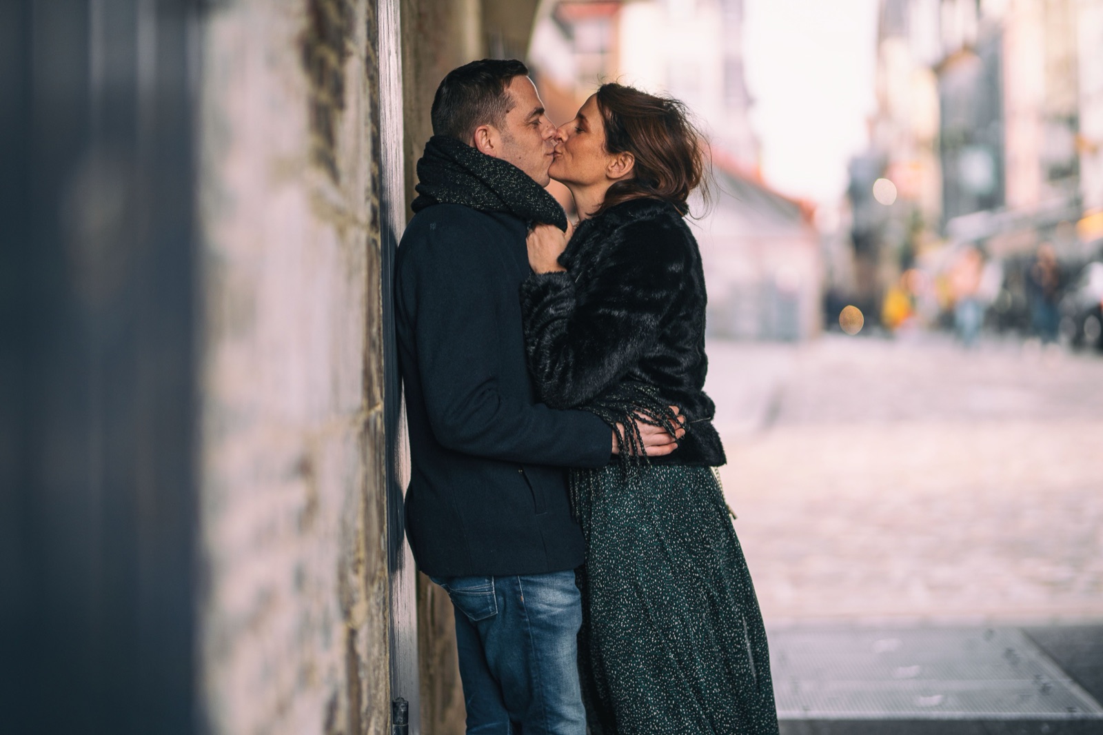 The couple kisses against a stone wall in a cobbled lane of old Honfleur — engagement session in Honfleur, Normandy — Arnaud Chapelle