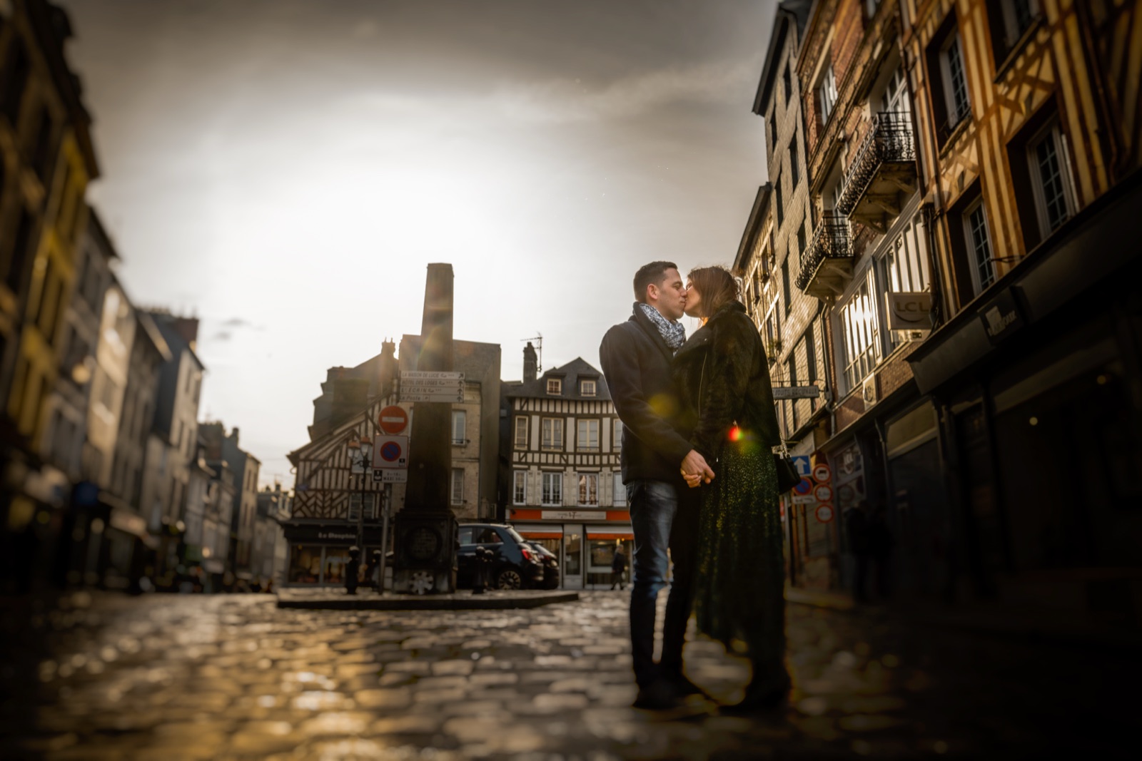 The couple kisses in the middle of a cobbled square in Honfleur, golden backlight on the half-timbered facades — engagement session in Normandy — Arnaud Chapelle