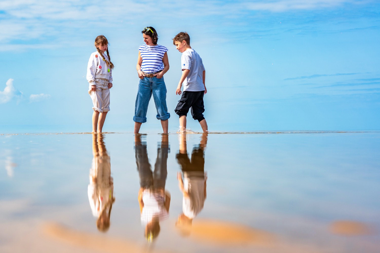 A mother and her two children walking barefoot on the wet sand at Omaha Beach, their reflection below them