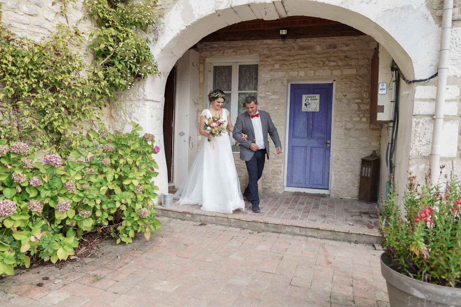 Les mariés sortent sous une arche de pierre fleurie d'hortensias — mariage au Cœur de Combray, Ernes — Arnaud Chapelle