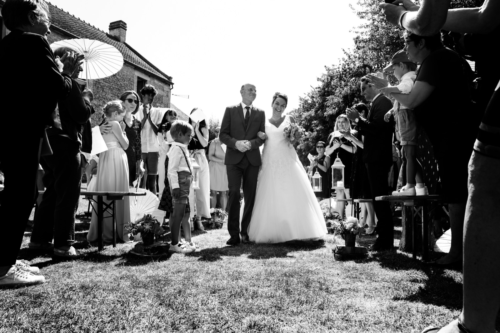 L'entrée de la mariée au bras de son père, sous les regards des invités, en noir et blanc — cérémonie au Domaine de Cauvicourt — Arnaud Chapelle